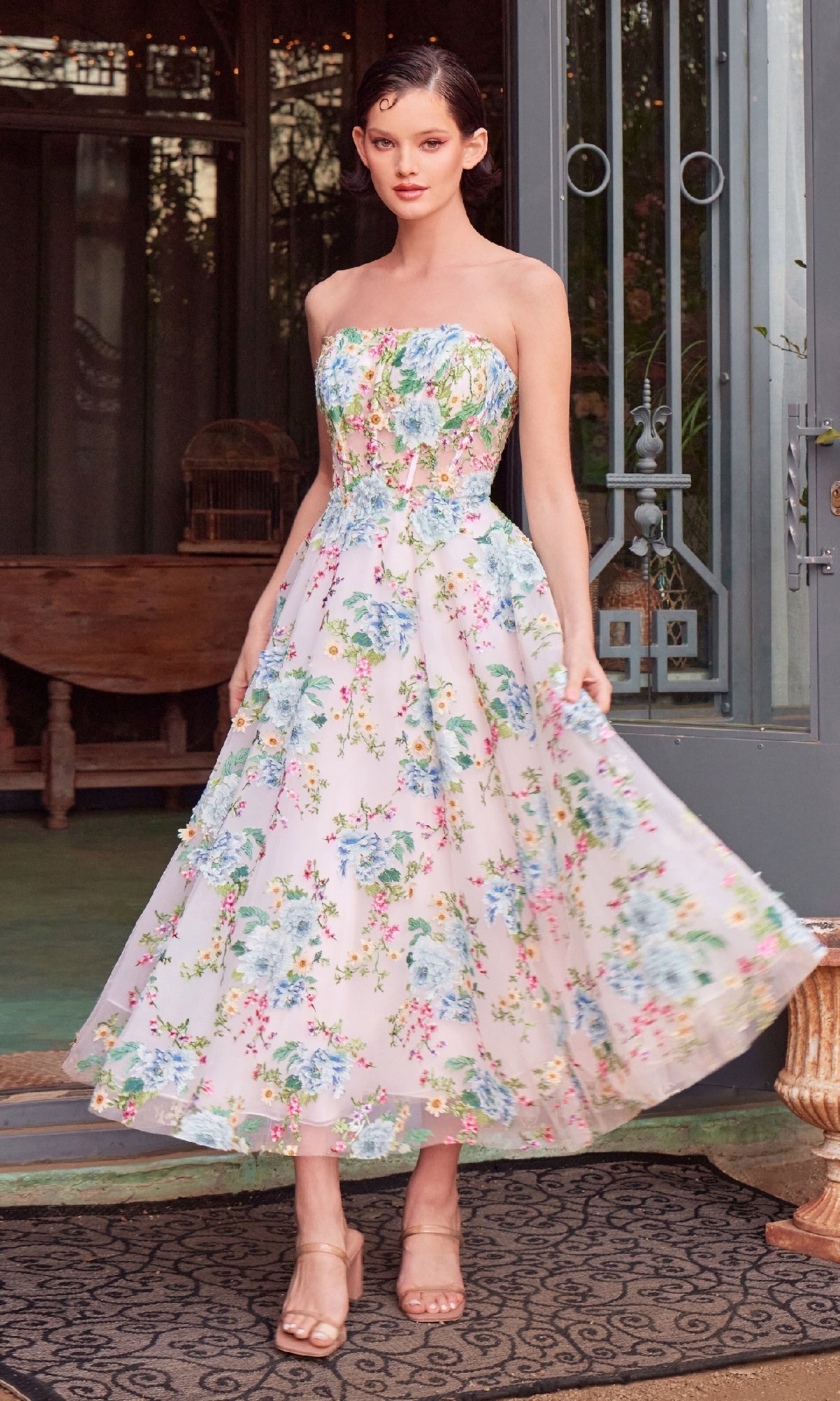 A woman in nude heels stands near a metal door and outdoor furniture, wearing the Andrea & Leo A1406 Floral Prom Dress with a strapless corset bodice.
